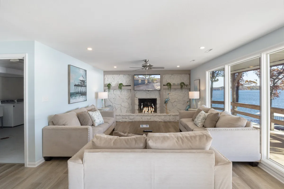 A spacious living room featuring comfortable sofas, a cozy fireplace, large windows overlooking the water, and tasteful artwork. The arrangement invites group gatherings while allowing for serene moments enjoying the view.