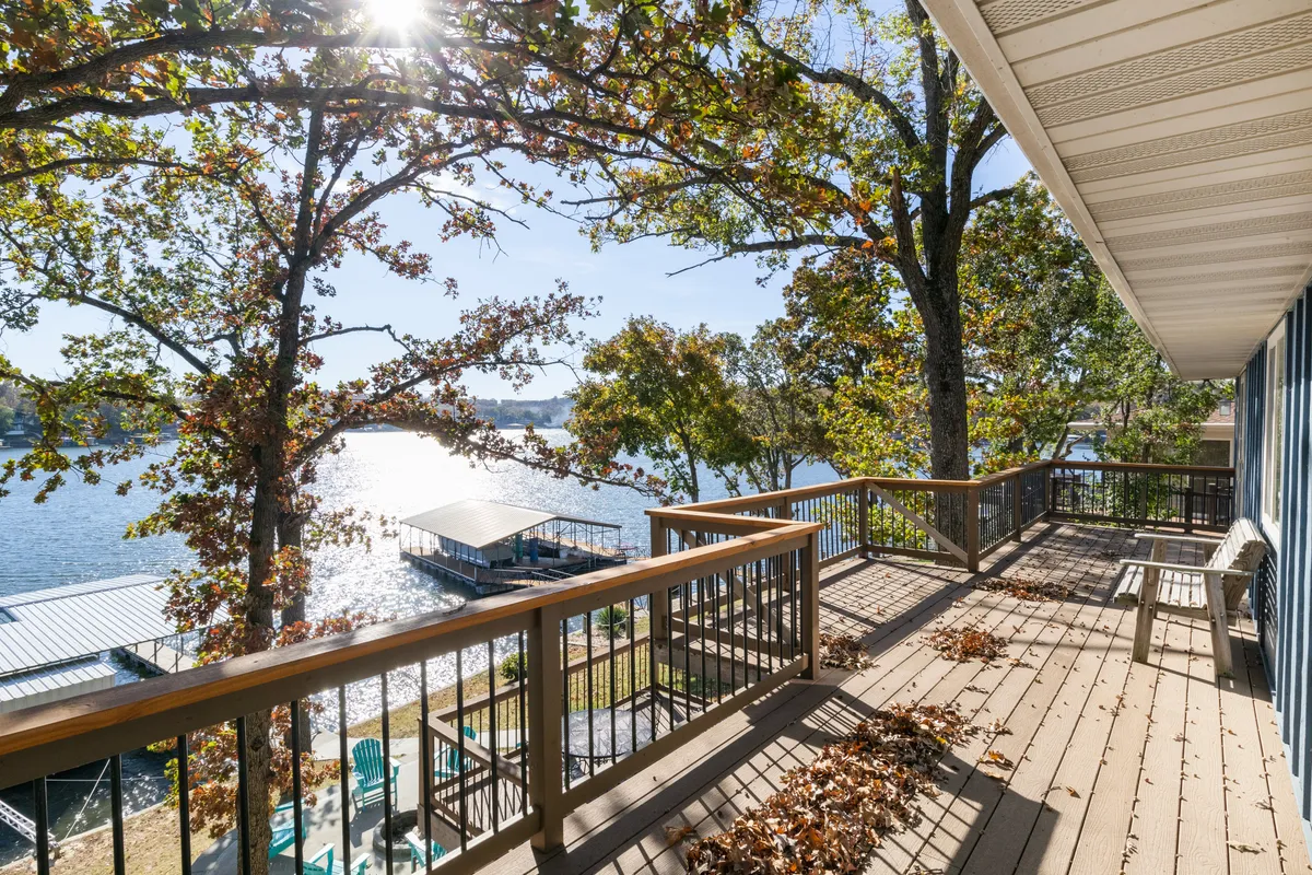 This image shows a peaceful lake view from the deck, with a deck railing in the foreground, surrounded by trees. The calm water stretches out with hints of autumn foliage, capturing the tranquility of lakeside living. The perspective from the deck invites viewers to imagine relaxing or gathering while overlooking the water.