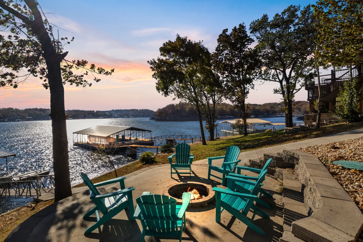 The image showcases the home's expansive lakeside deck with cozy Adirondack chairs encircling a glowing fire pit, perfectly positioned to take in sweeping sunset views over the tranquil lake. The sky is painted with warm evening colors, creating an inviting atmosphere ideal for gatherings and relaxation.