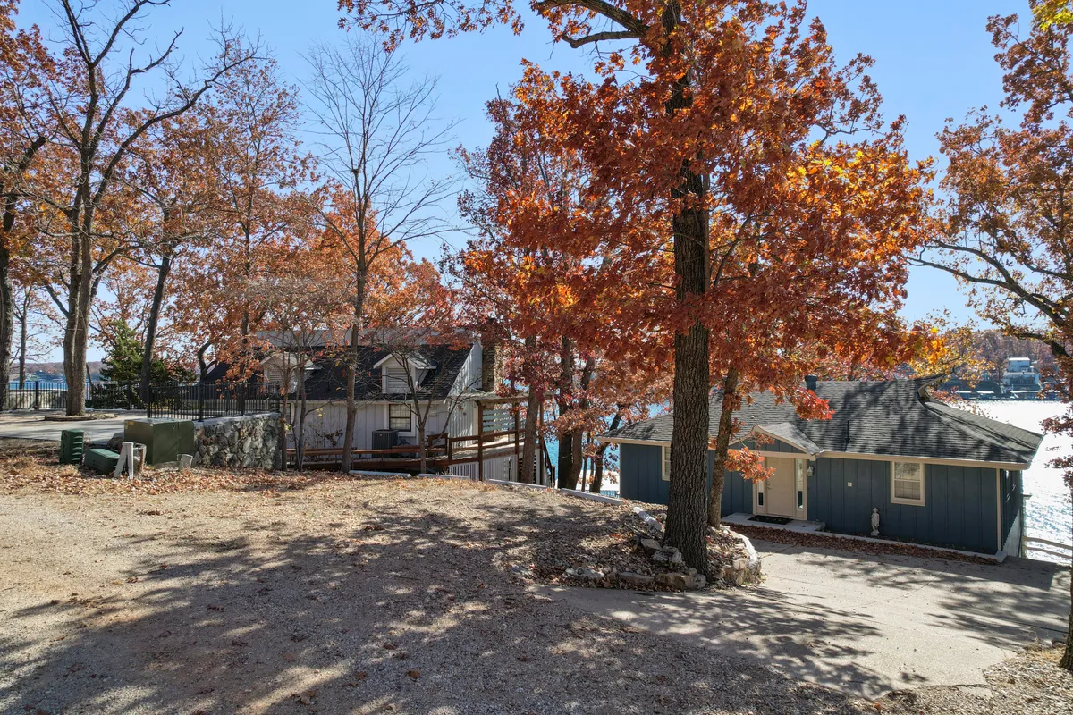 A serene lakeside scene featuring vibrant autumn trees, two distinct houses, and a clear water view. The setting is surrounded by mature foliage, highlighting the seasonal beauty of the Lake of the Ozarks region. The composition invites the viewer to imagine quiet nature walks, picnics, or simply relaxing outdoors.