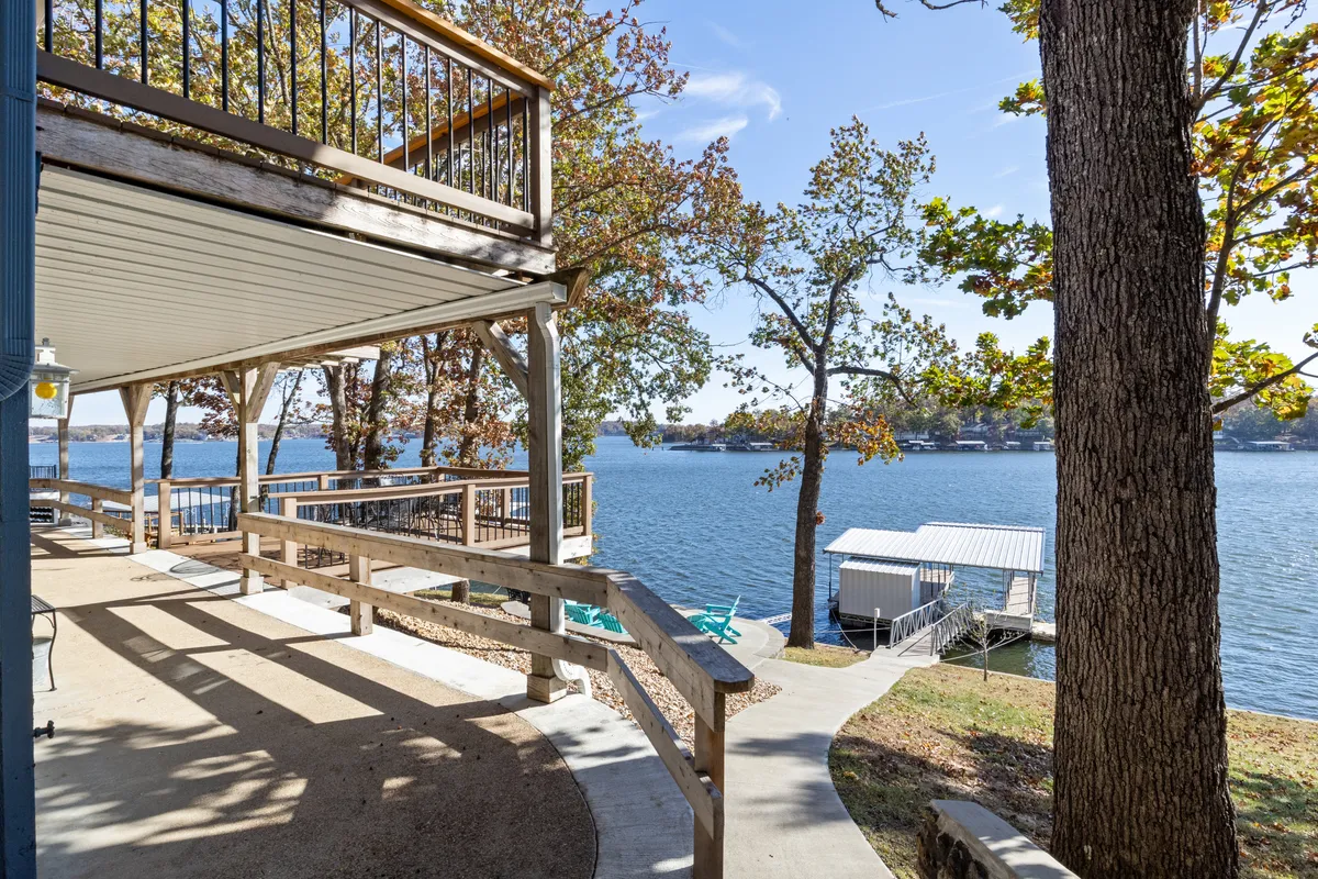 The photo features a wooden deck overlooking the lake, surrounded by trees and a boathouse. The setting exudes relaxation and offers a perfect spot for morning coffee, evening bonfires, or simply taking in the lakeside ambiance.