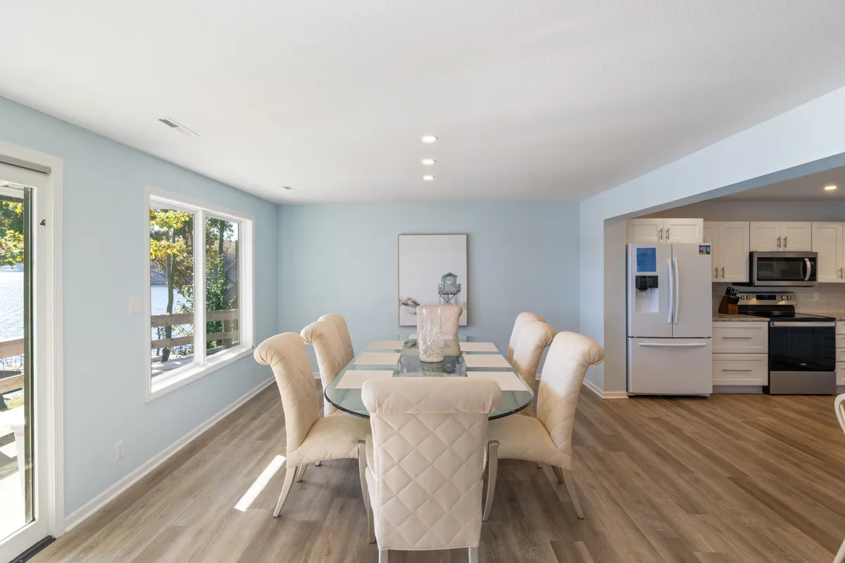 A dining area with elegant chairs and a table, set before large windows that overlook the lake. The adjacent kitchen area is visible, emphasizing the open-plan design and the beautiful, tranquil views that enhance every meal.