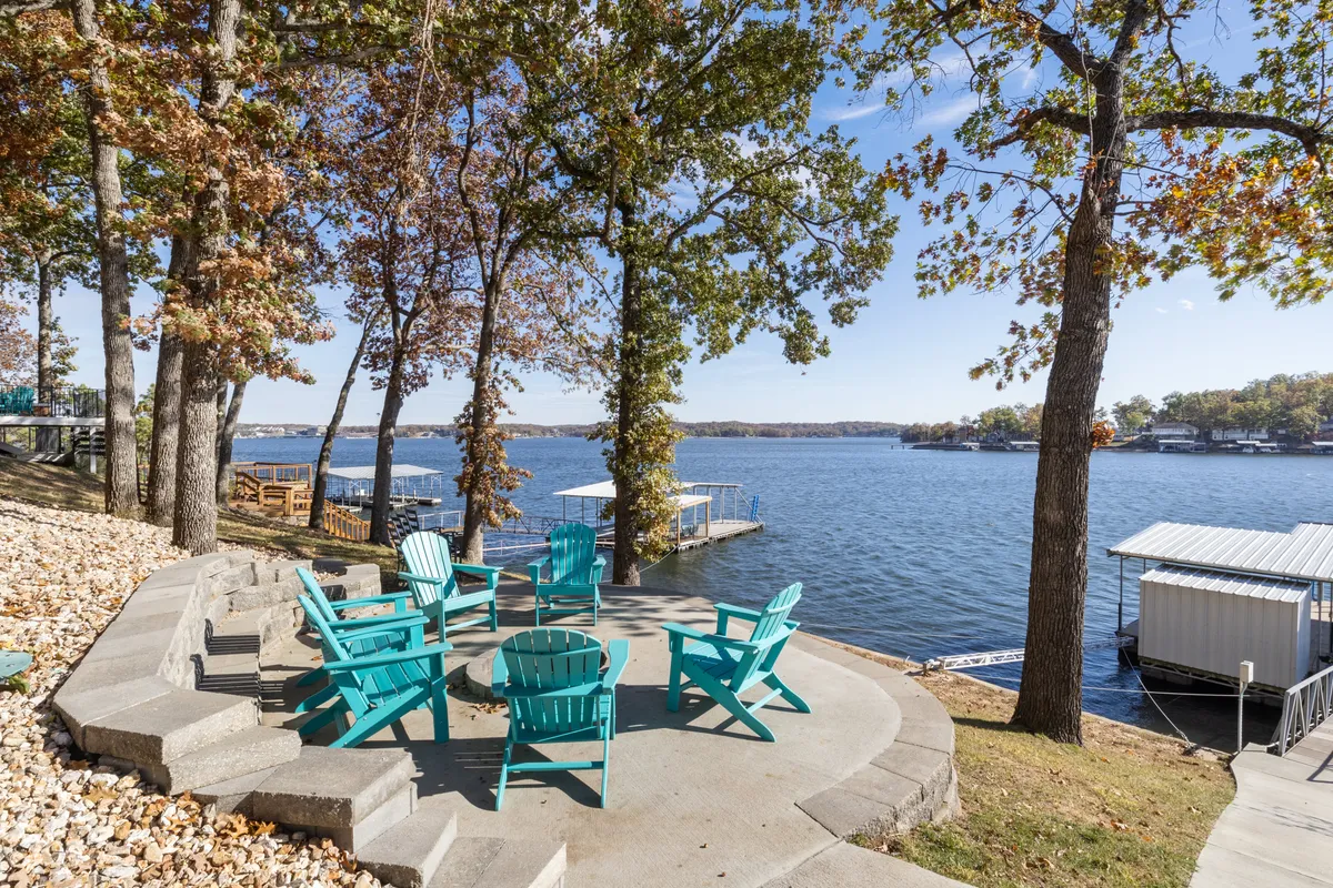 A set of bright turquoise Adirondack chairs faces the tranquil lake, surrounded by trees and set against a backdrop of calm water and sky. The setup evokes relaxation, socializing, and the enjoyment of lakeside leisure, with visible cues for gathering around a firepit or sharing stories by the water.