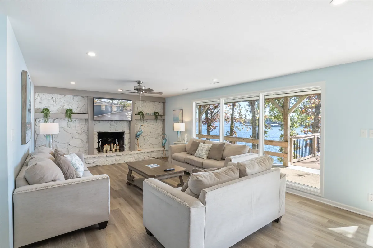 A spacious, inviting living room with a fireplace, large windows framing calming lake views, a coffee table, and plush seating arranged for family or group gatherings. Natural light floods the space, highlighting both comfort and scenic beauty.