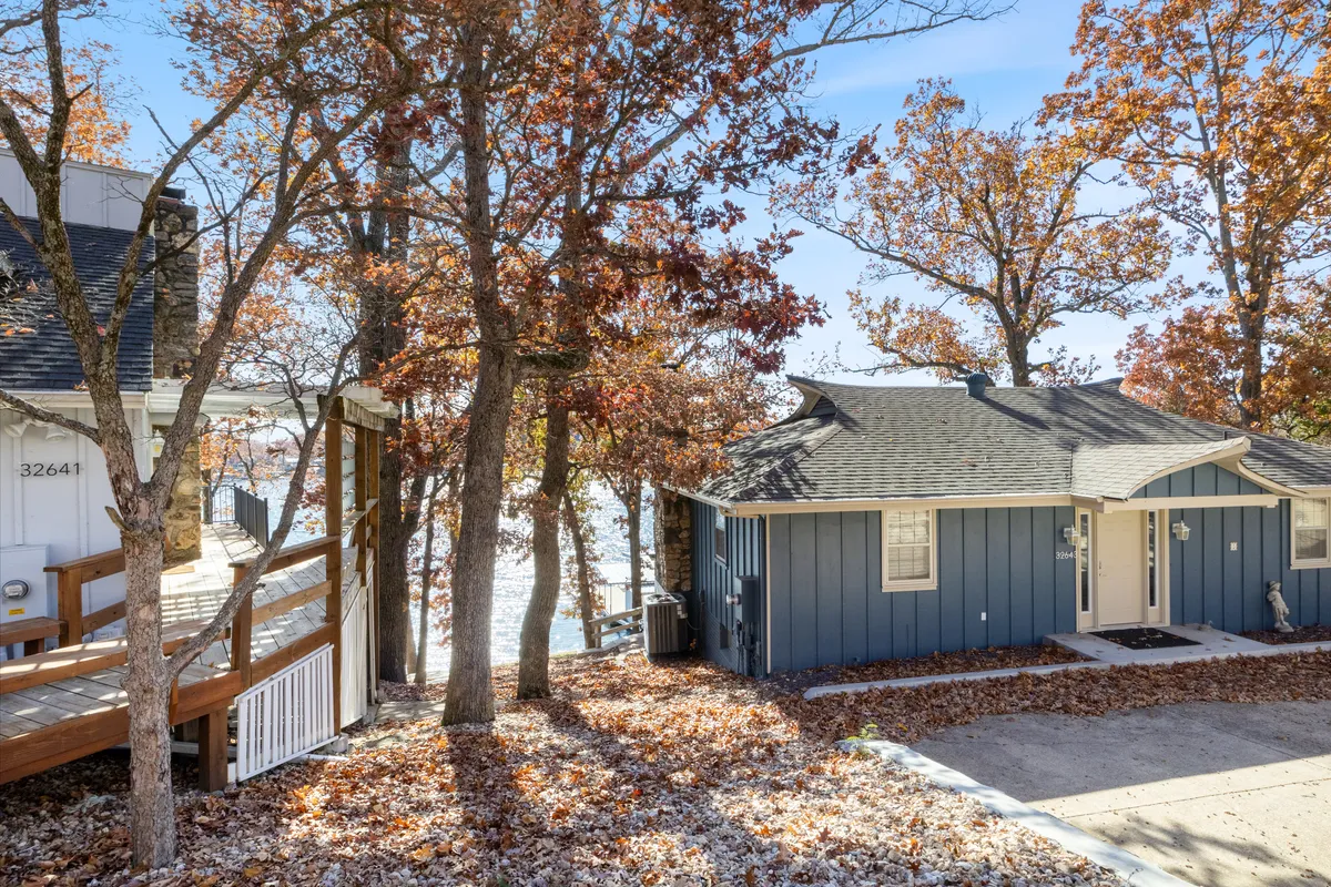 A serene exterior shot of a blue house framed by a tree canopy and overlooking the lake. The landscape is lush and green, hinting at multiple seasons of natural beauty. The scene evokes the feeling of returning to a peaceful, nature-filled retreat after a day of adventure.
