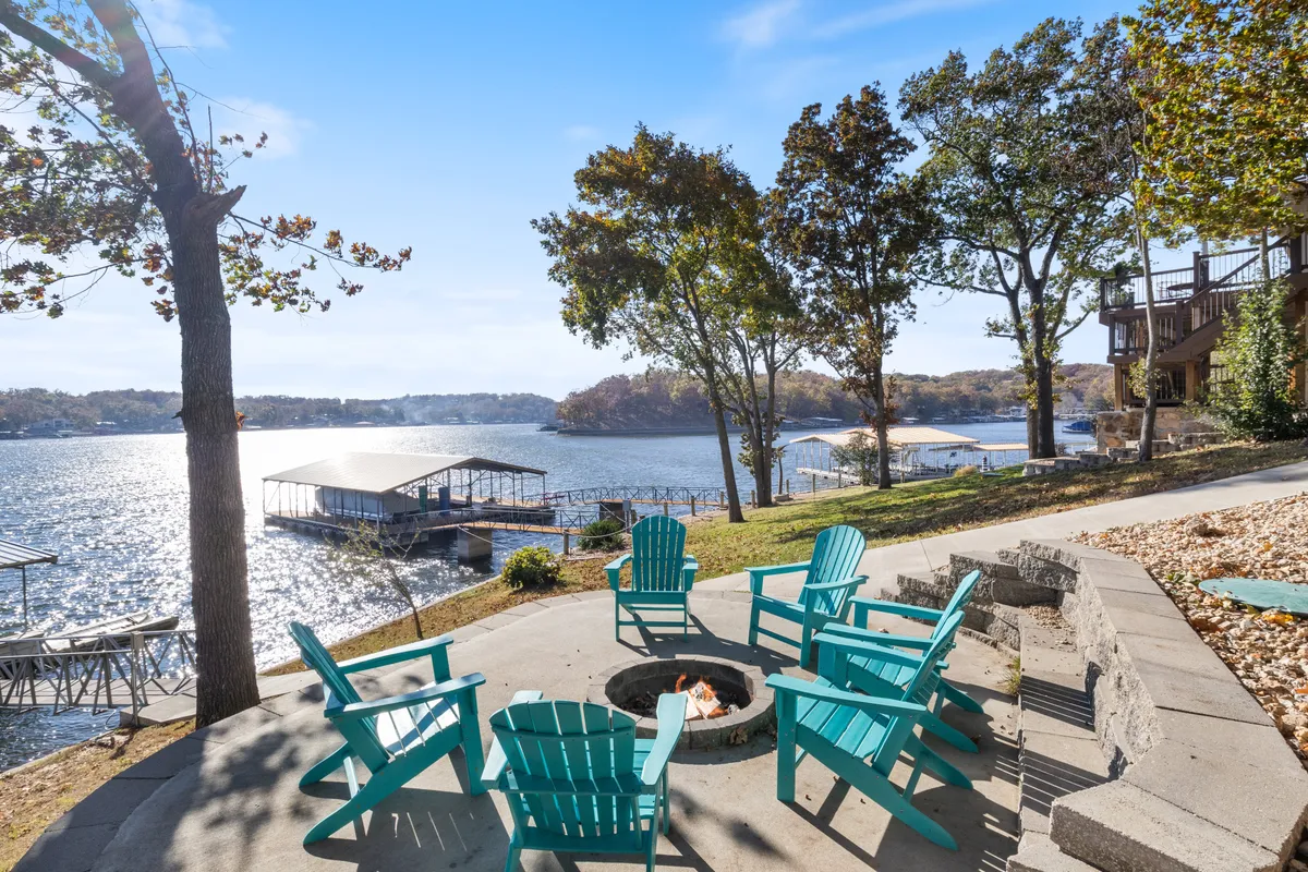 A cozy fire pit area featuring a circle of Adirondack chairs set on a deck or patio, all facing a beautiful lake view. The atmosphere is serene and communal, ideal for evening conversations, storytelling, and enjoying twilight by the water.