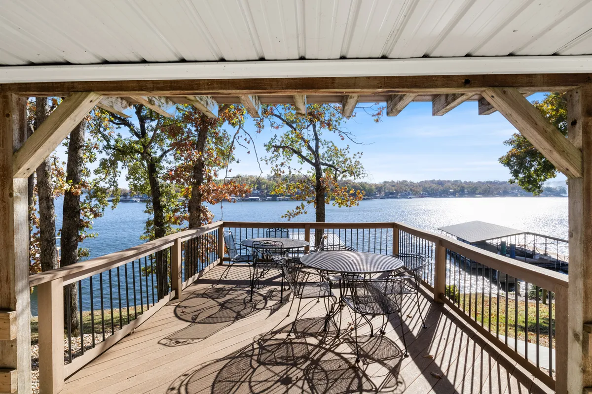A serene deck area furnished with outdoor seating and a dining table, overlooking a shimmering lake. Trees frame the scene, while the water view stretches out beyond the railing. The space is set up for both relaxation and al fresco dining, with inviting furniture and natural surroundings.