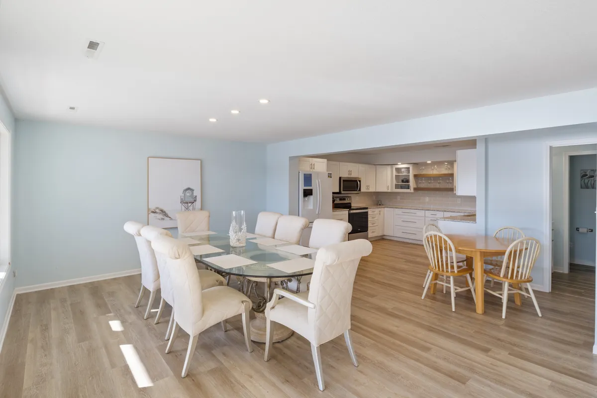 A bright, inviting open-plan space capturing a dining table with chairs and a modern kitchen area in the background. Elegant light fixtures add warmth, and the setup is clearly designed for group meals and easy interaction.