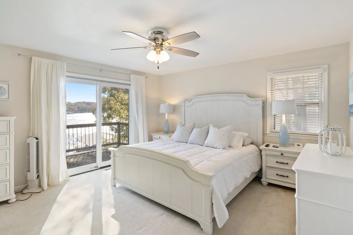 A tranquil bedroom featuring a cozy bed with soft linens, large windows that let in abundant natural light, a sliding door leading outside, and thoughtfully placed nightstands. The serene ambiance invites relaxation and restful sleep, and the view hints at the lakeside setting beyond.