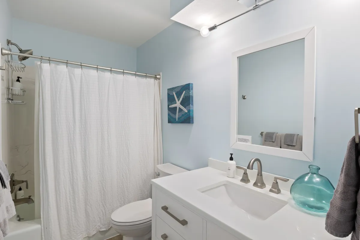 A modern, calming bathroom with a white shower curtain, blue starfish artwork adding a splash of color, a sleek vanity, and glass vessel. The space feels fresh, clean, and thoughtfully detailed, ideal for rejuvenation and self-care.